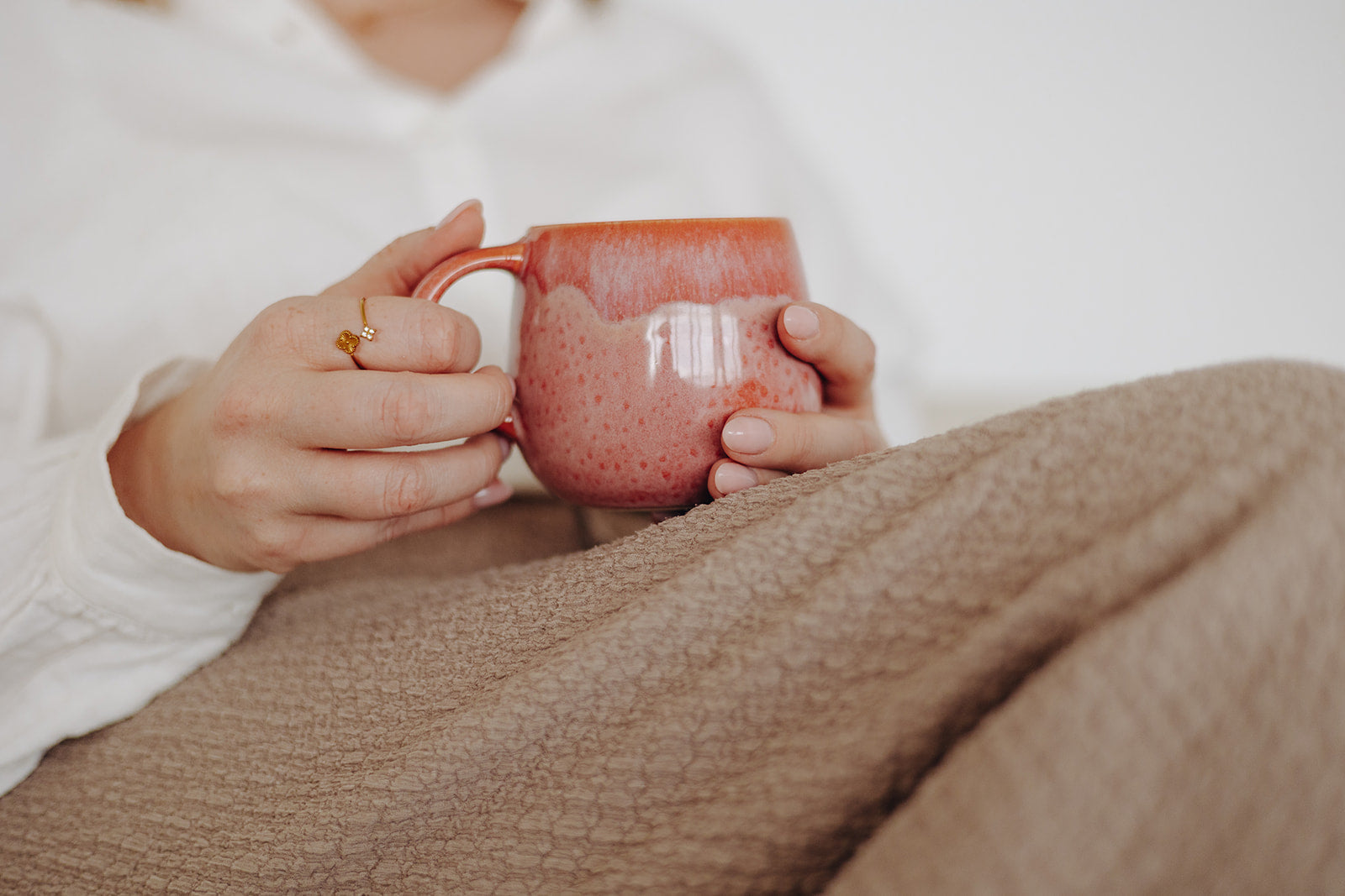 rosa rote Keramiktasse in Hand, ruhiger Kaffeemoment auf der Couch, verspielter Goldring am Finger